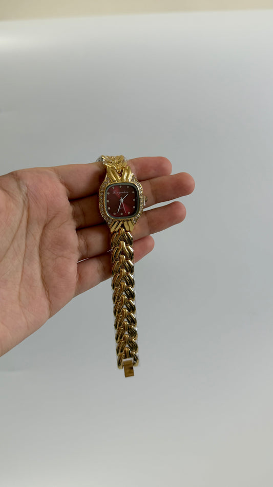 A gold-tone watch with a textured chain link bracelet and a dark red dial, held in a person's hand against a plain light gray background.